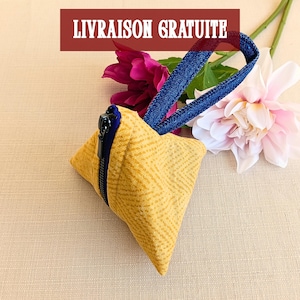 Puede incluir: Una bolsa de tela con forma de pirámide en color amarillo mostaza con un sutil patrón geométrico, que cuenta con una cremallera negra y una correa de muñeca de mezclilla azul oscuro. El texto LIVRAISON GRATUITE aparece en blanco sobre rojo.