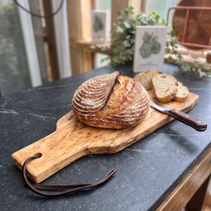 May include: A round loaf of sourdough bread with a scored crust sits on a long, light-colored wooden serving board with a dark leather hanging strap. A metal knife with a dark wooden handle rests next to several sliced pieces of bread.