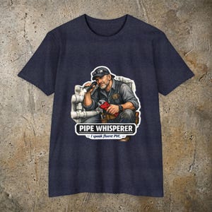 May include: A dark navy blue t-shirt featuring a graphic of a plumber holding a flashlight and a pipe wrench in front of white pipes. The text reads PIPE WHISPERER and I speak fluent PVC in white lettering below the illustration of the worker.