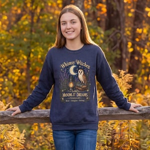 May include: A dark blue long-sleeve sweatshirt featuring a graphic print of an owl perched on books next to a lantern and a crescent moon. The text reads Whimsy Witches, Leni's MOONLIT DREAMS, Schattenwald, and Rest, Imagine, Belong in gold and white.