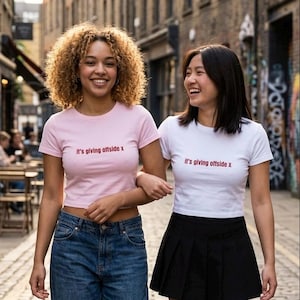 Puede incluir: Un top corto de manga corta rosa claro y un top corto de manga corta blanco, ambos con el texto "it's giving offside x" impreso en rojo en el pecho. El top rosa se combina con vaqueros azules y el top blanco con una falda negra.