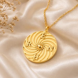 Könnte beinhalten: Eine goldfarbene, kreisförmige Anhängerkette mit einer zentralen, erhabenen Kugel, die von einem spiralförmigen Radialmuster umgeben ist. Die Buchstaben E, R, H und N sind an den Himmelsrichtungen auf die Scheibe geprägt.