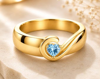 May include: A polished gold ring featuring a bypass-style band that curves to hold a single, small, light blue heart-shaped gemstone at the center. The metal band has a smooth, reflective finish and a continuous, flowing design.