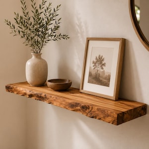 Könnte beinhalten: Ein schwebendes Wandregal aus Holz mit Naturkante. Auf dem Regal stehen eine beige Keramikvase mit grünen Blattzweigen, eine kleine dunkelbraune Holzschale und ein gerahmtes Bild mit einer schwarz-weißen Baumillustration.