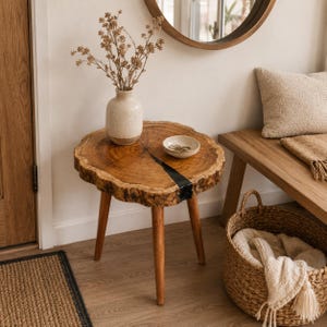 May include: A round side table featuring a natural wood slab top with a live edge and a black resin inlay, supported by three tapered wooden legs. A cream-colored ceramic vase with dried branches and a small white bowl sit on the table surface.