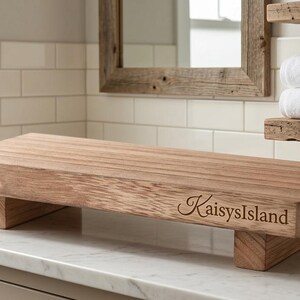 May include: A rectangular wooden riser with a natural light brown wood grain finish and four short block feet. The front edge of the wooden platform features the engraved text KaisysIsland in a cursive script font, showcasing a smooth surface.