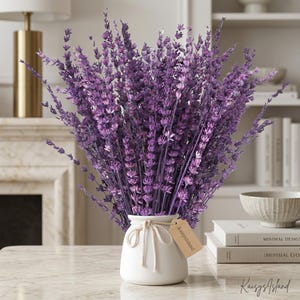 May include: A large bouquet of vibrant purple lavender stems arranged in a small, white ceramic vase. The vase is decorated with a simple white rope tie and a small, rectangular tan paper tag with the text KaisysIsland printed on it.