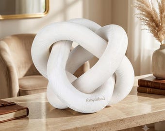 May include: A white wooden decorative knot sculpture with a visible wood grain texture. The object features an intricate, interlocking loop design and has the text KaisysIsland printed in a small, light font on one of the curved surfaces.