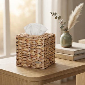May include: A square tissue box cover crafted from woven light brown natural fibers. The box features a small rectangular metal tag on the front side. A stack of white tissues protrudes from the top opening of the woven container.