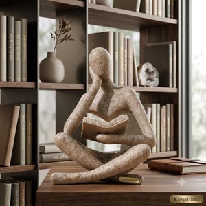 May include: A speckled beige abstract sculpture of a seated figure reading an open book. The figure rests its head on one hand while holding the book with the other. A small rectangular plaque with the text KaisysIsland is on the base.
