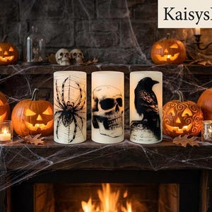 May include: Three cylindrical pillar candles featuring black graphic prints of a spider on a web, a human skull, and a raven perched on a tombstone. The candles are arranged on a mantelpiece alongside carved jack-o'-lanterns and small skulls.