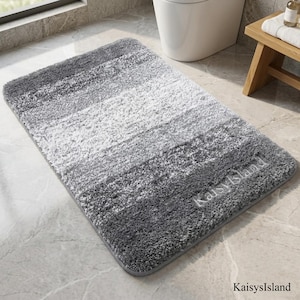 May include: A rectangular bath mat featuring a shaggy, high-pile texture with horizontal stripes in varying shades of gray and white. The text KaisysIsland is embroidered in a light gray thread on the bottom right corner of the mat.