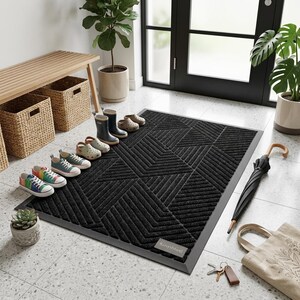 May include: A rectangular black doormat with a textured geometric pattern of raised lines and a small grey label in the corner. The mat is placed on a light-colored tiled floor near a wooden bench, several pairs of shoes, a black umbrella, and a tote bag.