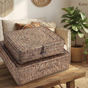 May include: Two rectangular woven seagrass storage baskets stacked on top of each other. The top basket features a hinged lid secured with a dark toggle closure. The brand name KaisysIsland is subtly embossed on the front of the bottom basket.