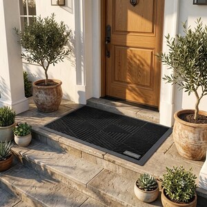May include: A rectangular black doormat featuring a textured geometric pattern of raised diagonal lines. The mat has a solid border and a small rectangular label in the bottom right corner, positioned on a stone porch in front of a wooden door.