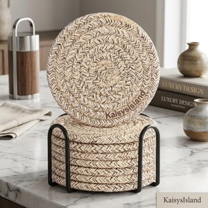 May include: A set of round, woven beige and brown coasters stacked in a black metal holder. The top coaster features the text KaisysIsland printed in a dark brown font. The coasters have a braided texture and a circular shape, designed for surface protection.