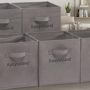 May include: Five square grey fabric storage bins with open tops and integrated handles on the front. Each bin features the text KaisysIsland printed in a dark grey font on the front panel. The bins have a subtle quilted texture on the exterior.