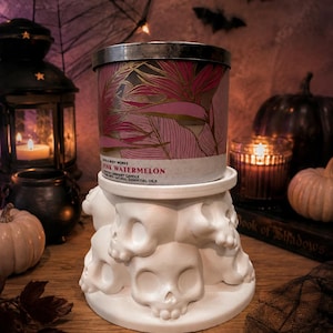 May include: A Bath & Body Works Pink Watermelon scented candle in a cylindrical jar with a pink and gold leaf pattern, resting on a white candle holder base sculpted with multiple human skulls arranged in a circular formation.