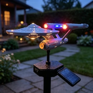 May include: A white starship model mounted on a black solar-powered garden stake. The spacecraft features a saucer section, two engine nacelles with glowing red and blue lights, and illuminated windows. The surface is covered in small water droplets.