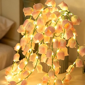 Puede incluir: Una guirnalda de luces con flores artificiales en forma de tulipán de pétalos rosa suave y bombillas LED de luz blanca cálida. Las flores están dispuestas a lo largo de finos cables verdes, creando un efecto de cascada con múltiples pétalos brillantes.