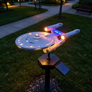 May include: A white starship model mounted on a black post with a solar panel. The spacecraft features a circular saucer section, two engine nacelles with glowing orange lights, and blue illuminated accents. The ship is illuminated from within.
