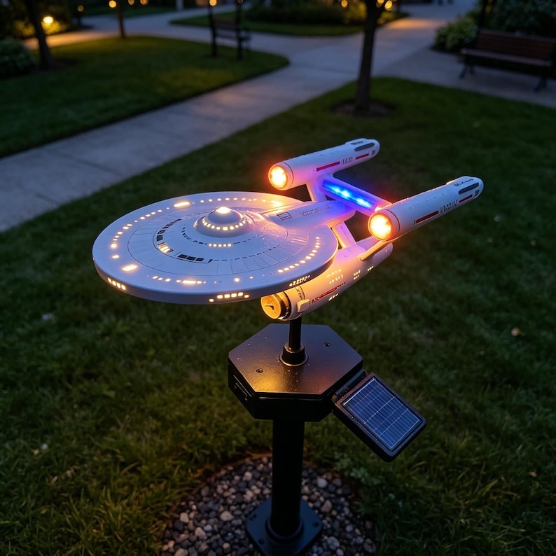 May include: A white starship model mounted on a black post with a solar panel. The spacecraft features a circular saucer section, two engine nacelles with glowing orange lights, and blue illuminated accents. The ship is illuminated from within.