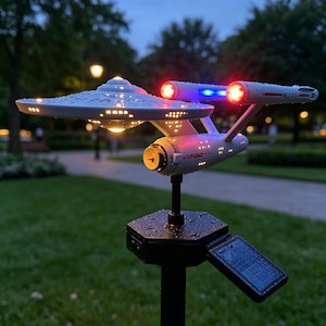 May include: A white Starship Enterprise model solar garden light featuring a saucer section, two nacelles with glowing red and blue lights, and a black hexagonal base with a small solar panel, all covered in light water droplets.