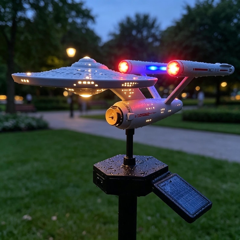 May include: A white Starship Enterprise model solar garden light featuring a saucer section, two nacelles with glowing red and blue lights, and a black hexagonal base with a small solar panel, all covered in light water droplets.