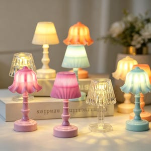 May include: A collection of small, battery-operated table lamps in various colors and shapes, including fluted shades in pink, blue, and orange, and clear faceted designs. Each lamp features a slender base and emits a soft, warm glow.