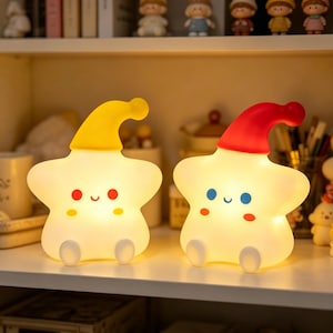 May include: Two star-shaped night lights with glowing white bodies and smiling faces. One star wears a yellow nightcap with red cheeks, while the other wears a red nightcap with blue eyes and orange cheeks. Both have small white feet extending forward.