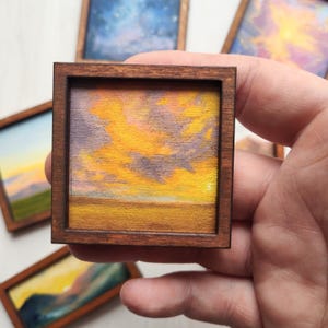 May include: A small square painting in a dark brown wooden frame depicting a sunset with vibrant yellow and orange clouds over a golden horizon. The artwork features textured brushstrokes and is surrounded by other similar miniature landscape paintings.