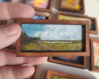 May include: A miniature rectangular oil painting in a dark wood frame, depicting a landscape with a green field, a distant horizon line, and a bright blue sky with thick, textured white clouds applied with visible brushstrokes.