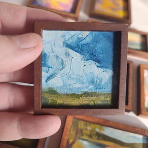 May include: A miniature square oil painting in a dark brown wooden frame, depicting a landscape with a textured blue sky featuring white clouds above a green field. The artwork is part of a collection of small framed landscape paintings.