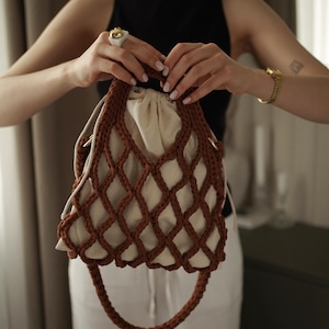 May include: A brown crocheted net handbag featuring a drawstring closure with a cream-colored fabric lining. The bag has a thick, braided brown handle and a matching long shoulder strap, showcasing a structured, open-weave design.