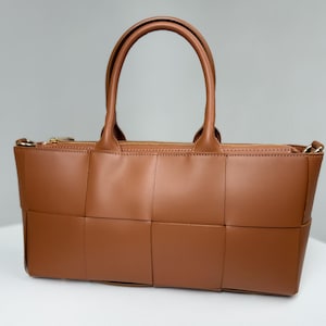 Può includere: Una borsa rettangolare marrone caratterizzata da una struttura in similpelle intrecciata con ampi pannelli quadrati. La borsa presenta due manici superiori, una chiusura a zip color oro sul bordo superiore e anelli metallici laterali.