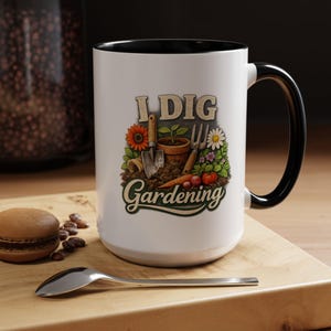 May include: A white ceramic mug with a black interior and handle, featuring a colorful graphic of gardening tools, a potted plant, flowers, and vegetables with the text I DIG Gardening printed in a stylized font on the front surface.