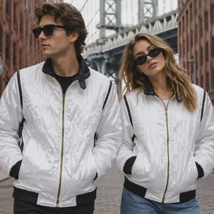 Puede incluir: Dos chaquetas bomber blancas idénticas con patrón acolchado de diamantes, cremallera frontal dorada de longitud completa, cuello acanalado negro, puños acanalados negros y paneles laterales verticales negros. Las chaquetas tienen un corte estructurado.