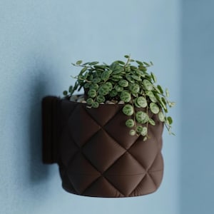 Puede incluir: Una maceta de pared de color marrón oscuro con un exterior acolchado de patrón de diamantes. El recipiente cilíndrico contiene una planta verde colgante con hojas pequeñas, redondas y variegadas sobre guijarros claros.