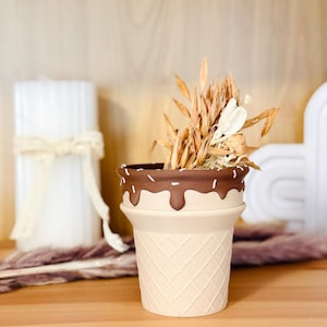Op de afbeelding: Een keramische plantenpot in de vorm van een ijshoorntje met een beige wafelstructuur en een donkerbruine rand met een druipend chocoladeglazuurontwerp met kleine witte spikkels, gevuld met een bosje gedroogde beige en witte plantenstengels.