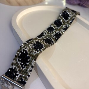 May include: A wide beaded bracelet featuring a repeating geometric pattern in black, olive green, and silver seed beads. The bracelet is finished with a silver-toned metal clasp and an adjustable chain extension with a small heart-shaped charm.