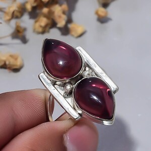 Könnte beinhalten: Ein Silberring mit zwei tropfenförmigen, tiefvioletten Cabochon-Steinen, die nebeneinander angeordnet sind. Das Metallband hat eine polierte Oberfläche mit einem strukturellen Design, das die beiden glatten, abgerundeten Edelsteine einrahmt.