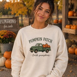 Puede incluir: Una sudadera de cuello redondo color crema con un gráfico de una camioneta vintage verde que transporta calabazas naranjas. El texto sobre la camioneta dice PUMPKIN PATCH en verde oscuro, y el texto debajo dice fresh pumpkins.