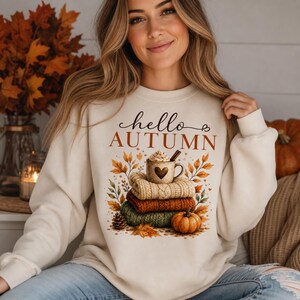 Puede incluir: Una sudadera de cuello redondo color crema con el texto "hello AUTUMN" en letras marrones. El gráfico muestra una pila de tres jerséis doblados, una taza con un corazón, una pequeña calabaza y hojas de otoño de colores.