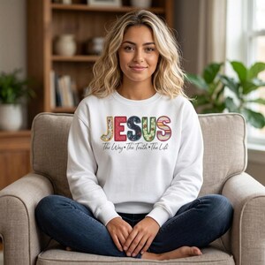 Puede incluir: Una sudadera blanca de manga larga con la palabra JESUS en letras grandes, coloridas y con diversos patrones en el pecho. Debajo, el texto dice The Way + The Truth + The Life en una fuente cursiva sencilla y de color oscuro.