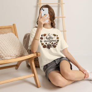 Puede incluir: Una camiseta de manga corta color blanco roto con un estampado gráfico que incluye el texto "hello autumn" rodeado de ilustraciones de hojas de otoño, una luna creciente, bellotas, setas, una calabaza y una taza humeante.