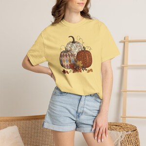 Puede incluir: Una camiseta de manga corta de color amarillo pálido con un estampado gráfico de tres calabazas. Las calabazas muestran diferentes patrones, incluyendo diseños de rombos, florales y filigranas, acentuados con hojas de otoño y bellotas.