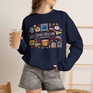 Puede incluir: Una sudadera azul marino de cuello redondo con un estampado gráfico que incluye el texto Stars Hollow Connecticut. El diseño presenta ilustraciones de un letrero de restaurante, un cenador, libros, una taza de café, un dónut y hojas de otoño.