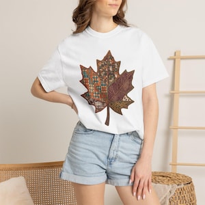 Puede incluir: Una camiseta blanca de manga corta con un gráfico central grande de una hoja de arce. El diseño de la hoja está dividido en secciones que muestran un mosaico de varios patrones, incluyendo formas geométricas y pequeños animales.