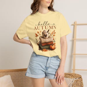 Puede incluir: Una camiseta de manga corta de color amarillo pálido con un estampado gráfico de una taza con un corazón, jerséis de punto apilados y una calabaza. El texto "hello AUTUMN" aparece impreso encima en una fuente marrón oscura.