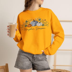 Puede incluir: Una sudadera de manga larga de color naranja brillante con un estampado gráfico de tres calabazas en tonos naranja, azul y blanco, rodeadas de enredaderas y hojas verdes, con el texto Pumpkin Season escrito en una fuente cursiva negra.